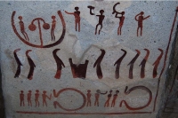 Petroglyph with a top row of people with musical instruments, a middle row of three otters on the left and five on the right with a cauldron between the groups, and people in the bottom row arranged in two groups of four, each facing an omega-shaped figure on its side, open towards them (some kind of enclosure?)